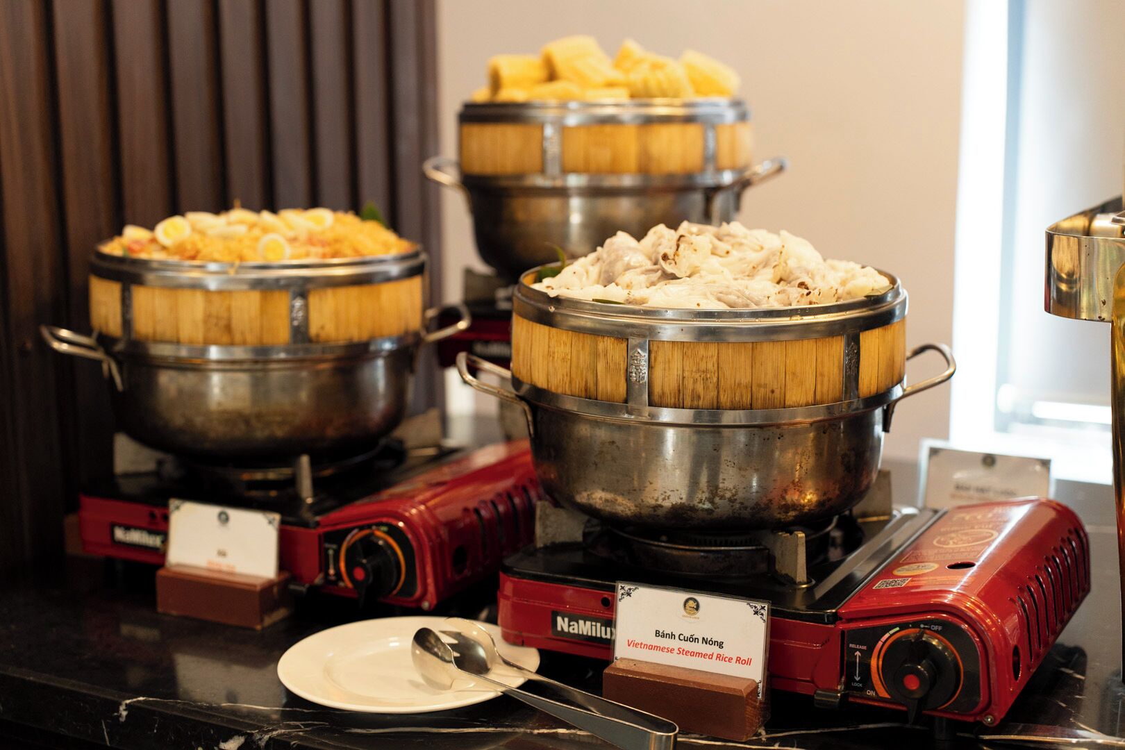 A buffet with various dishes, including Vietnamese steamed rice rolls, served in wooden bowls on electric stoves.