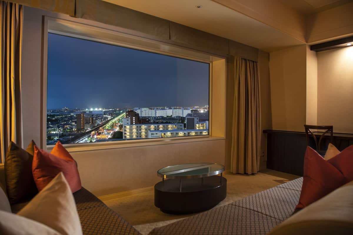 Habitación de hotel con un ventanal grande que ofrece vistas nocturnas de la ciudad, una mesa de centro redonda de vidrio y una zona de estar con cojines.