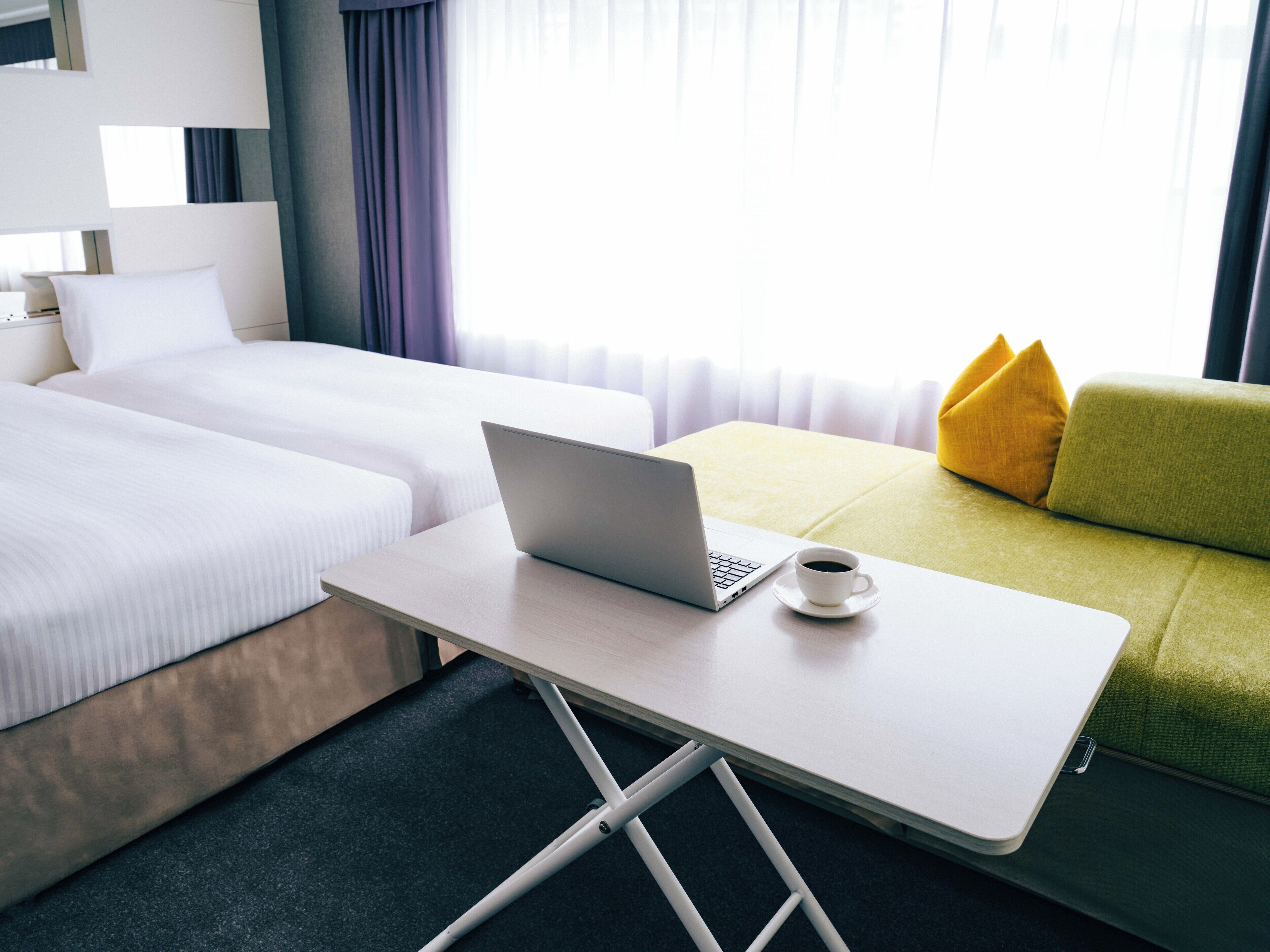 A hotel room with two beds, a desk with a laptop and a cup, and a green sofa with yellow cushions.