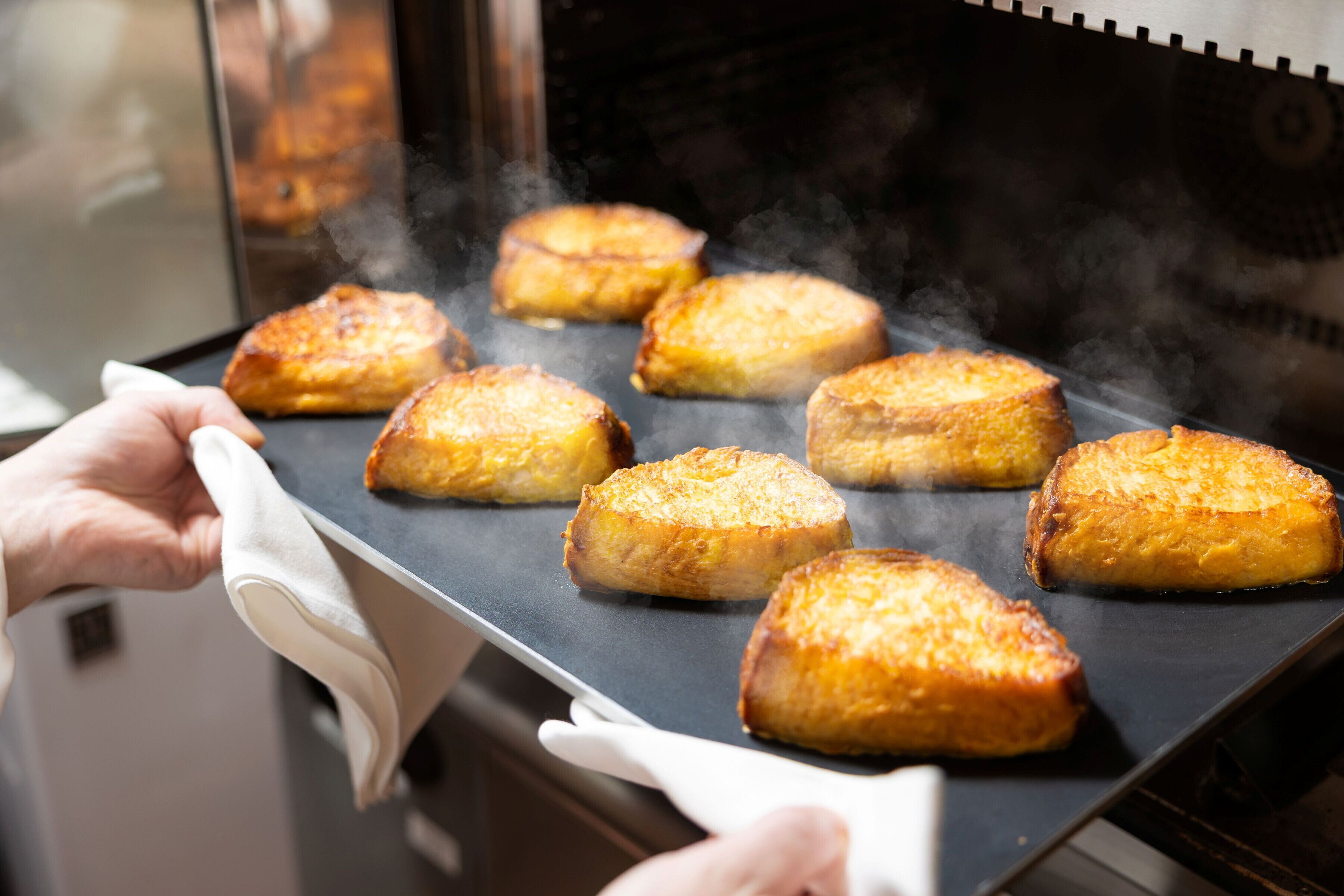 Eine Person hält eine Platte mit mehreren goldbraunen Backwaren, vermutlich Brot, die auf einem Grill gebacken werden.