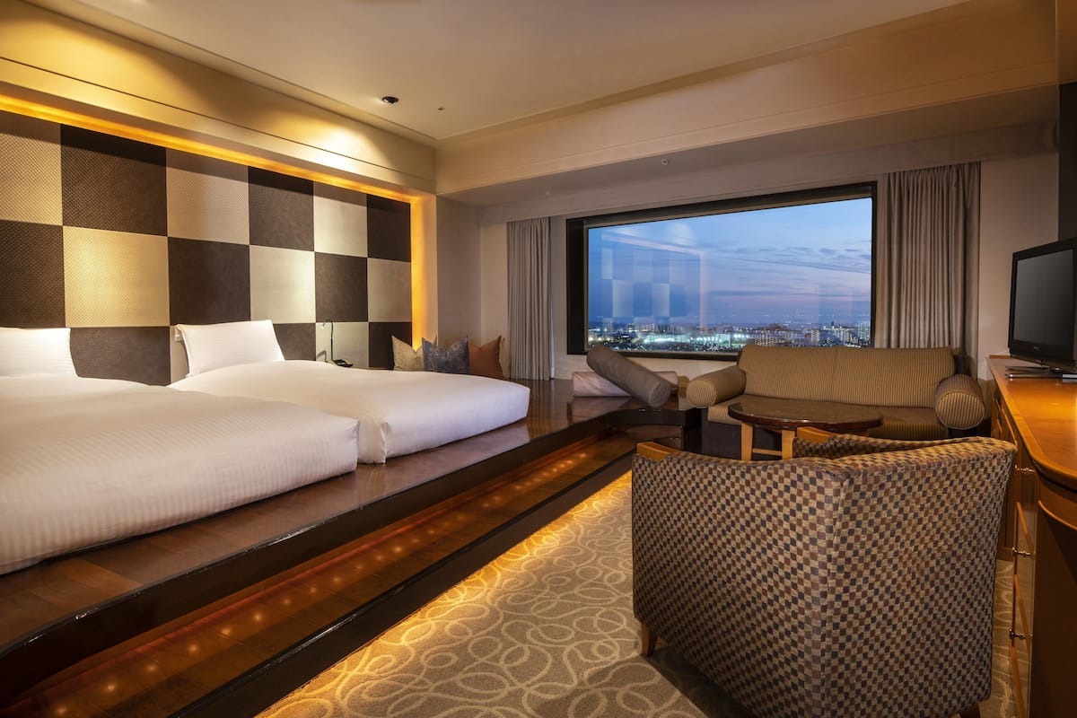Habitación de hotel con dos camas, un sofá, una silla, un televisor y un ventanal con vistas a la ciudad.