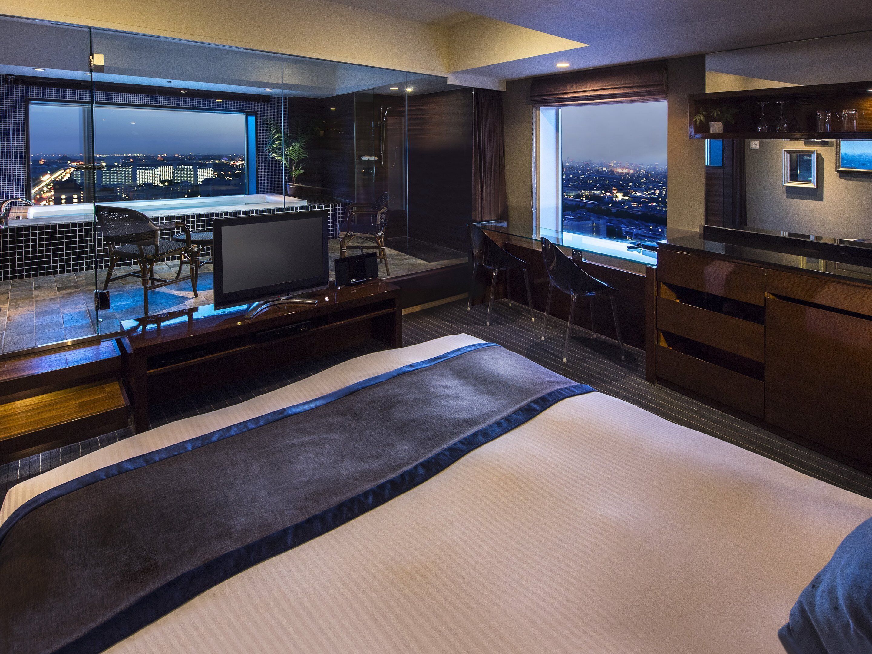 A modern hotel room with a large bed, a desk with a chair, a view of the cityscape, and a balcony with a seating area.