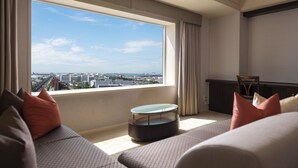 Ein modernes Hotelzimmer mit großem Fenster und Blick auf die Stadt, einem runden Couchtisch und einer Couch mit gemusterten Kissen.