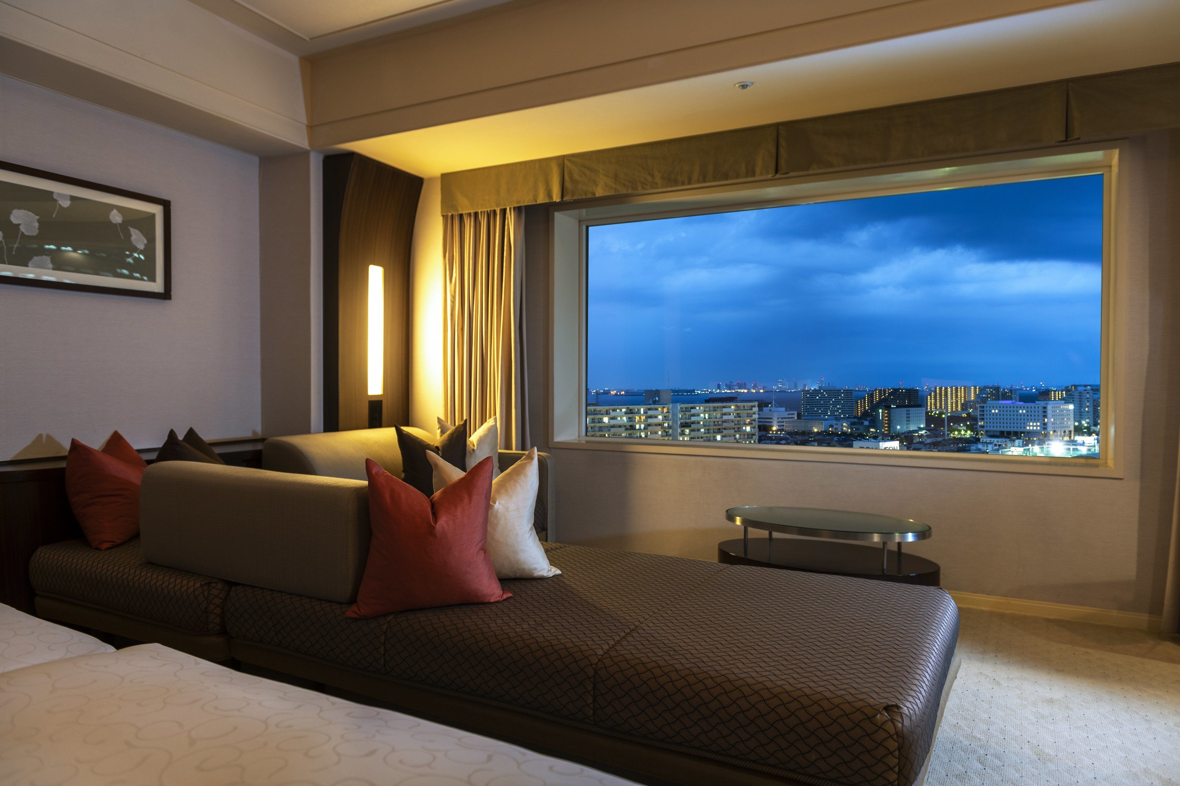 A hotel room with a large window offering a city view, a sofa with patterned cushions, and a bedside table.
