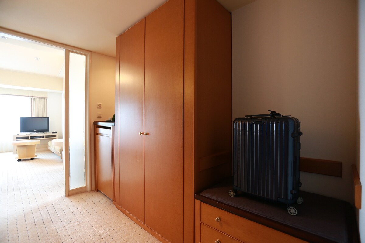 Ein Zimmer mit Kleiderschrank, Schreibtisch, Fernseher und Koffer.