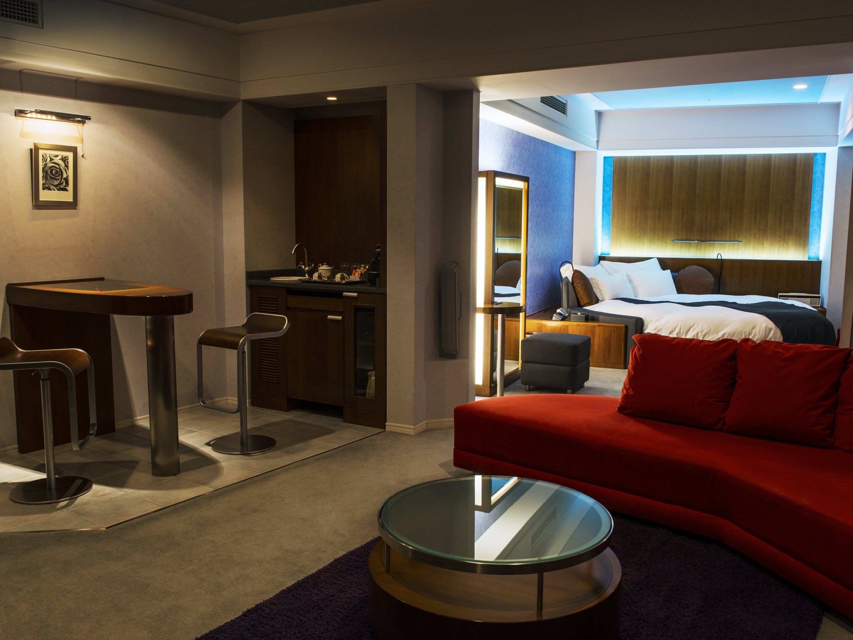 A hotel room with a bed, a red sofa, a glass coffee table, a desk with two chairs, a kitchenette, and a bathroom.