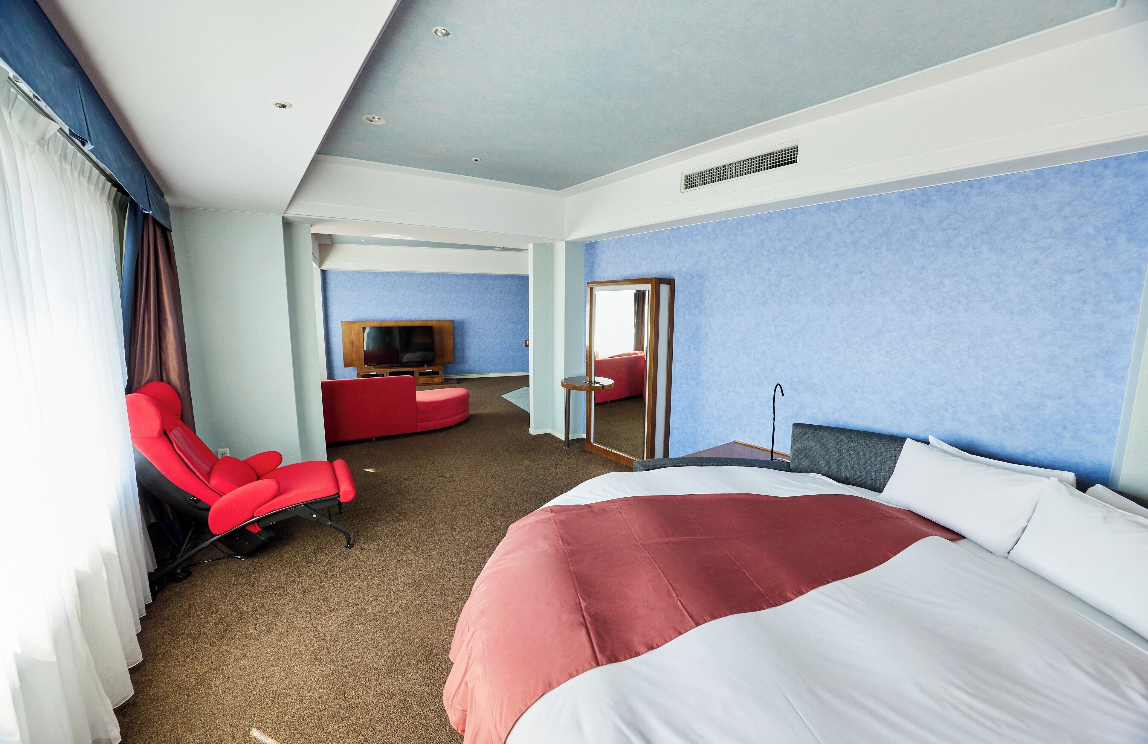 Una habitación de hotel con una cama grande, un sillón rojo, un televisor y un espejo.