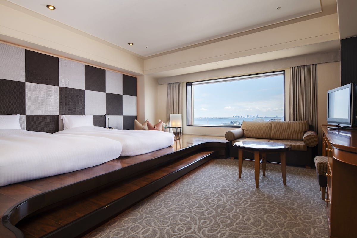 Habitación de hotel con dos camas, un sofá, televisión y un amplio ventanal con vistas a la ciudad.