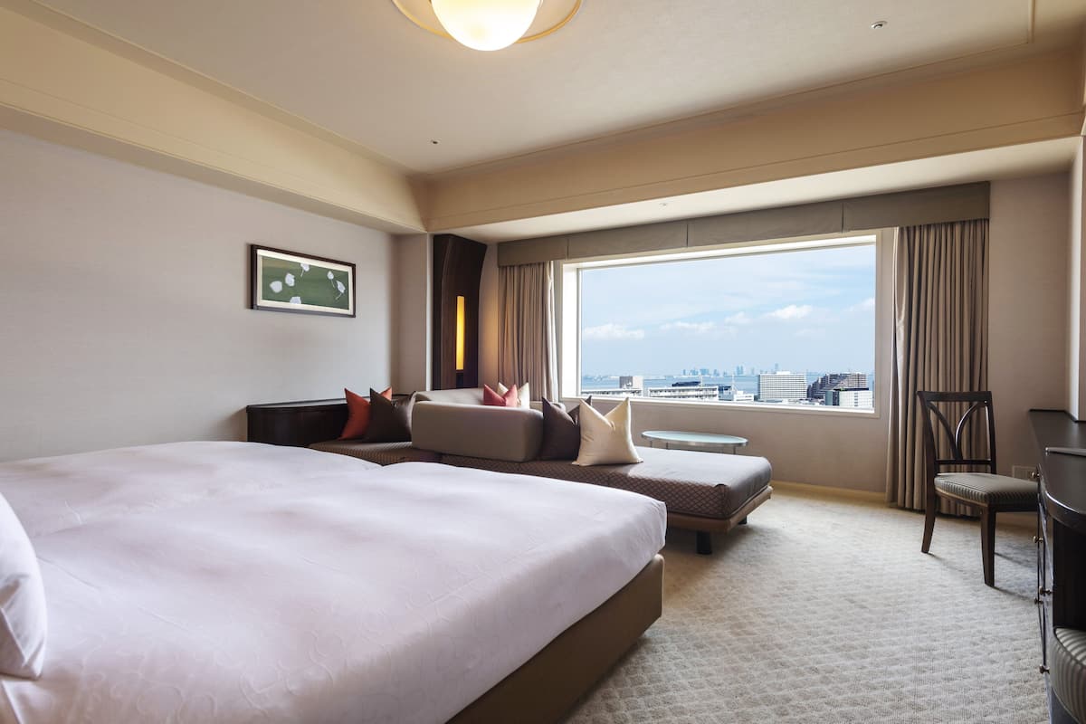 Habitación de hotel con dos camas grandes, un sofá, un escritorio y una silla. Amplia ventana con vistas a la ciudad.