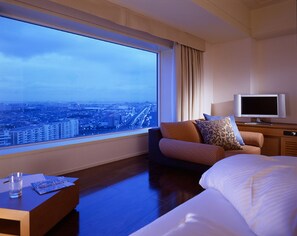 A hotel room with a large window, a bed, a TV, and a seating area with cushions.