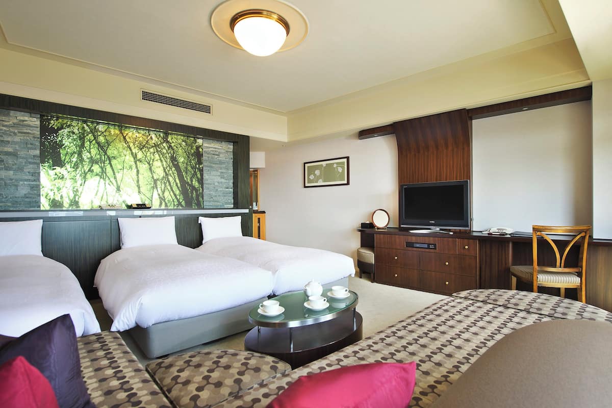 Habitación de hotel con dos camas, televisión, escritorio y una ventana grande con vistas a árboles.