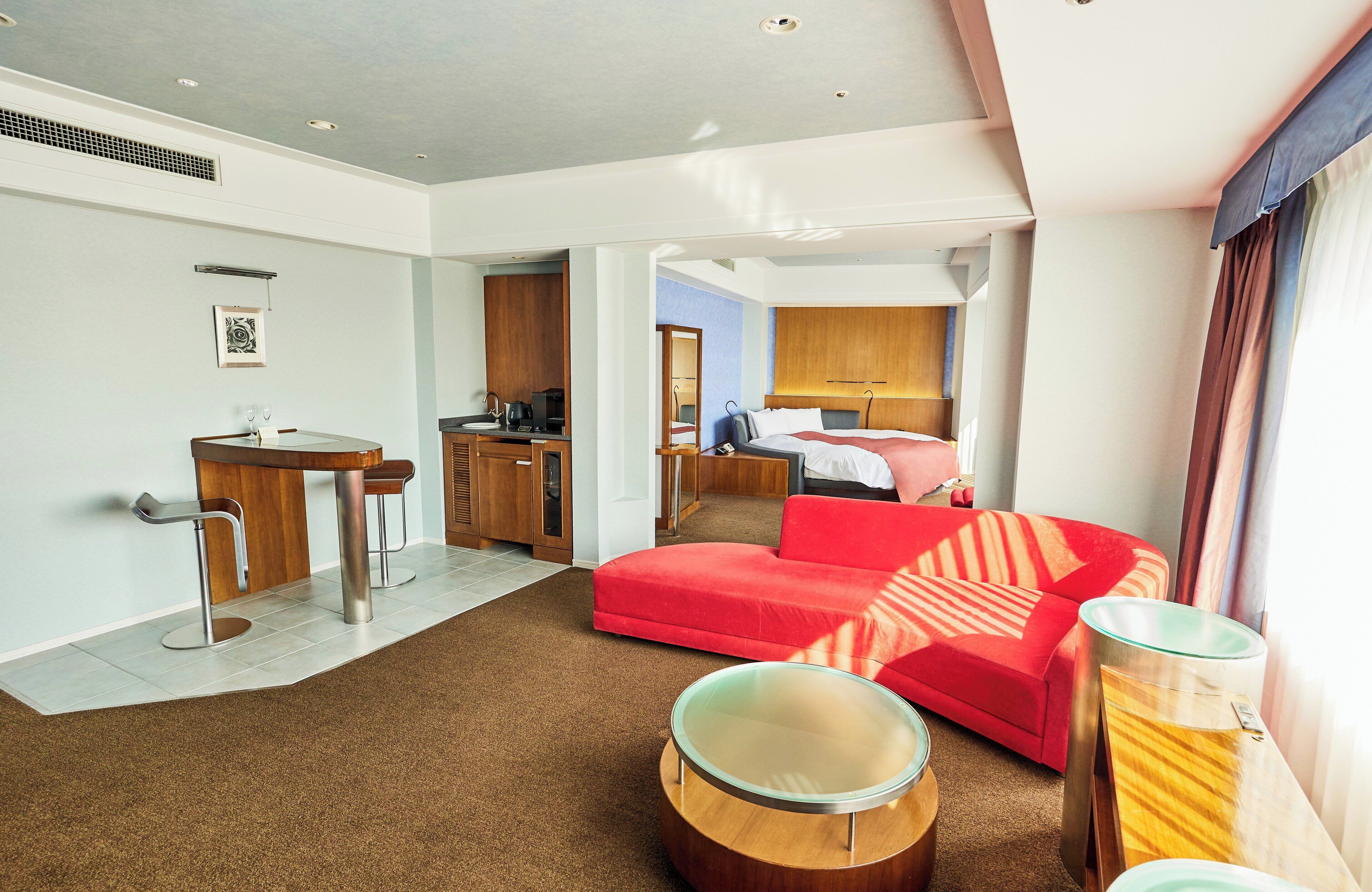 A modern hotel room with a red sofa, a glass coffee table, a dining area with a bar, and a separate bedroom with a bed and a nightstand.