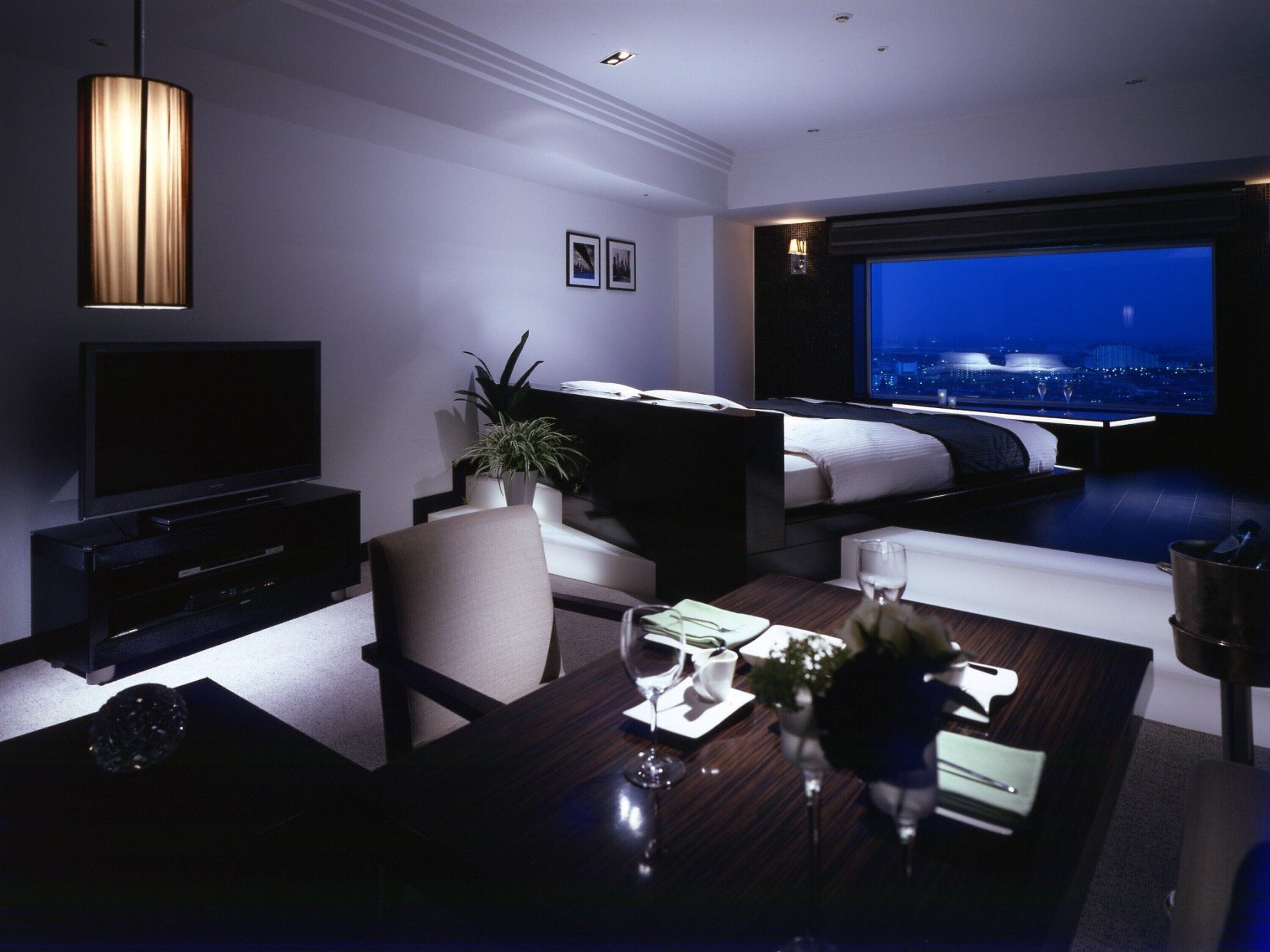 A modern hotel room with a flat-screen TV, a sofa, a dining table with a vase of flowers, and a city view through the window.