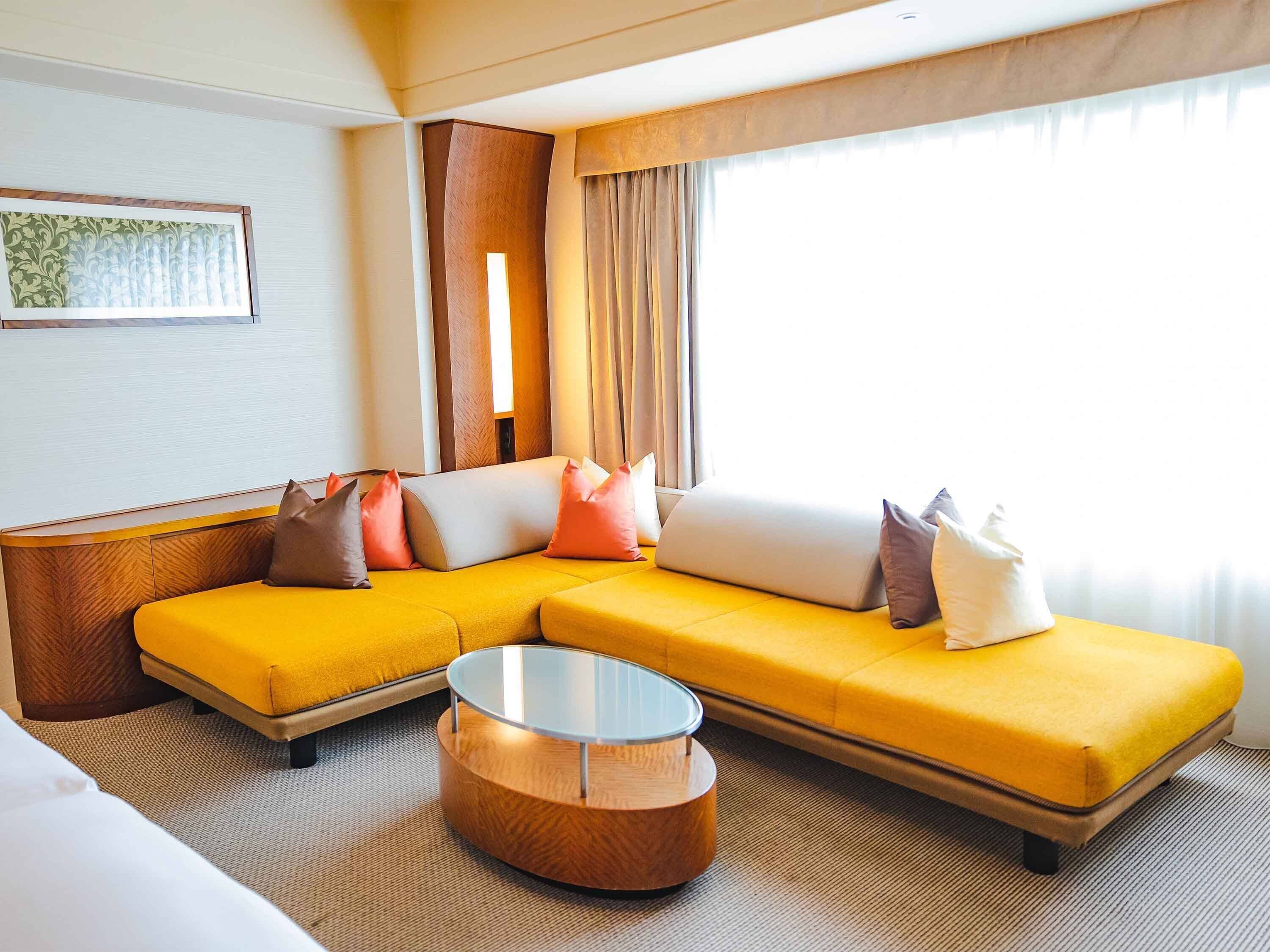 A modern hotel room with a yellow sectional sofa, a glass coffee table, and a large window with curtains.
