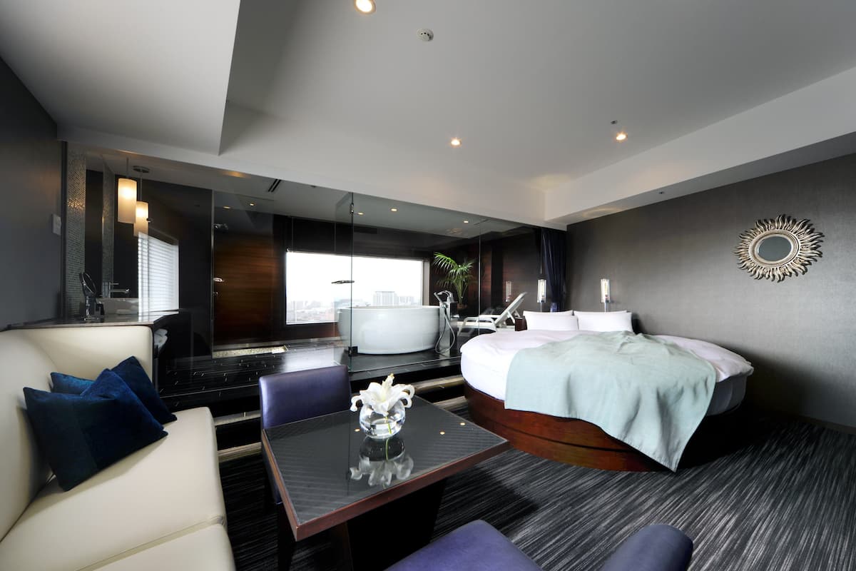 A modern hotel room with a large bed, a sitting area with a table, and a view of the city through a large window.