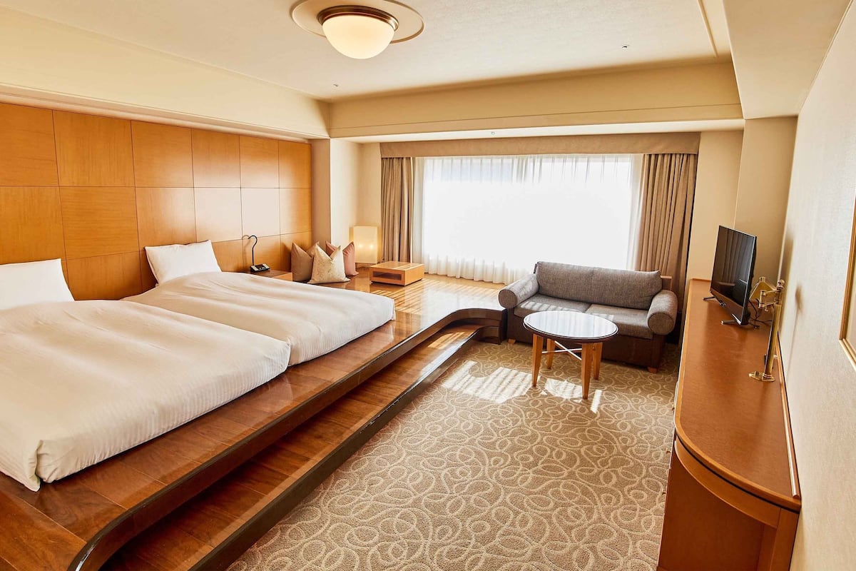 Habitación de hotel con dos camas, un sofá, una mesita, televisión y un ventanal grande con cortinas.