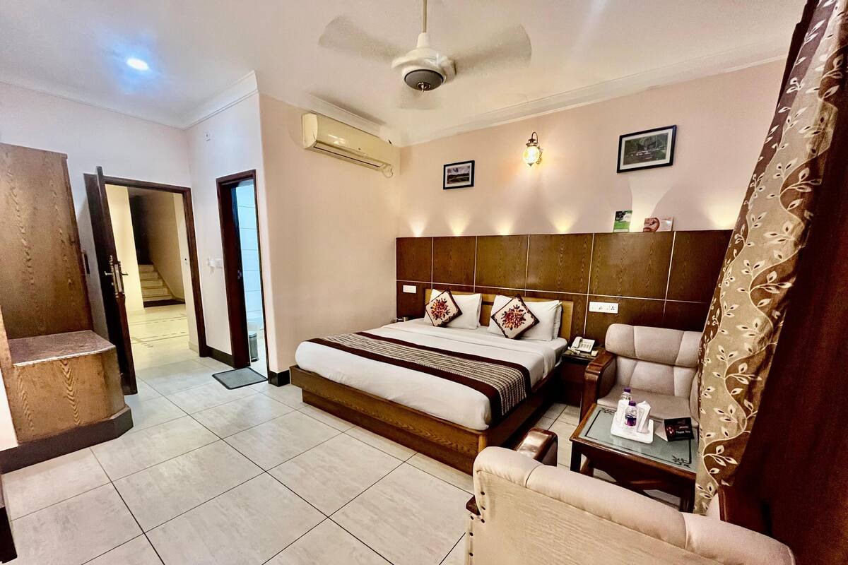 Habitación de hotel con una cama grande, una silla, una mesita de noche, un ventilador de techo, un equipo de aire acondicionado y una puerta que da a otra habitación.