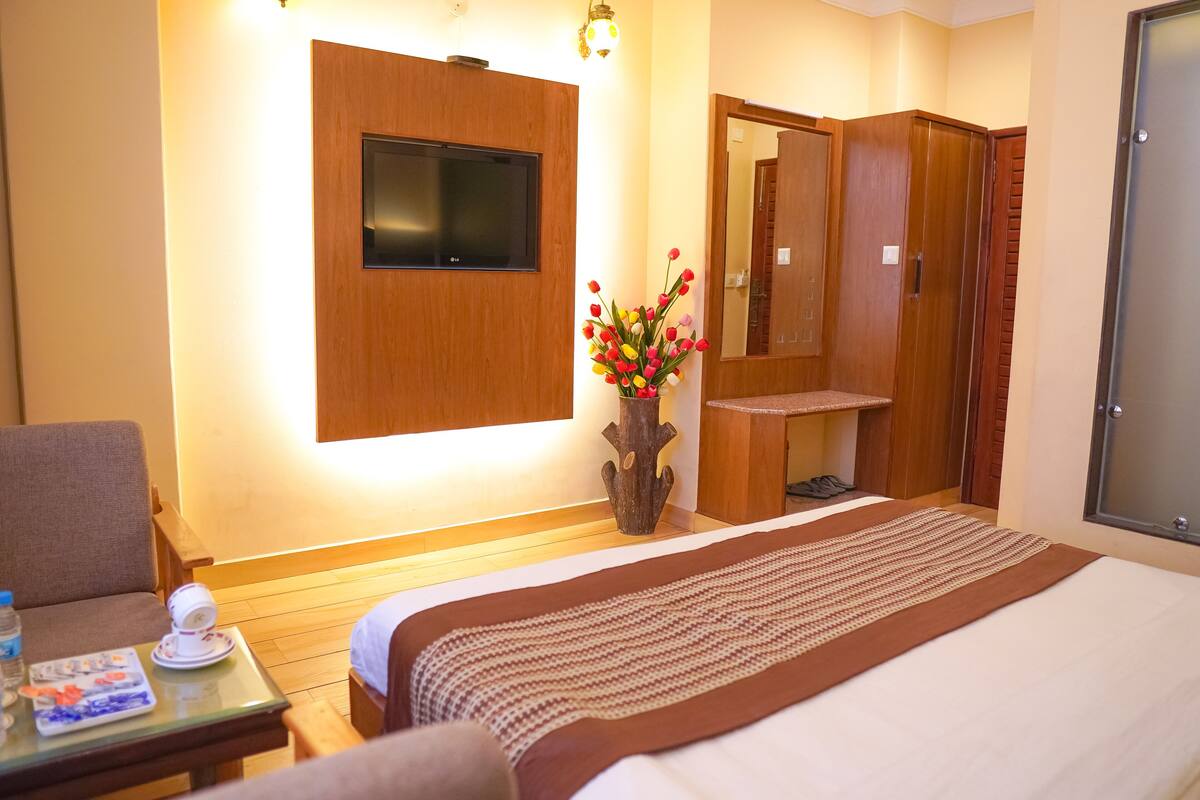 Habitación de hotel con una cama, un televisor montado en la pared, un armario de madera, una mesita con un jarrón de flores y una silla.