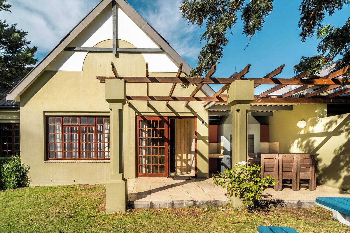 A house with a triangular roof, a wooden pergola, a front door, and a small patio area with a table and chairs.