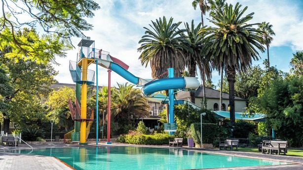 A swimming pool with a water slide, palm trees, and benches. - ATKV Goudini Spa (Rawsonville)