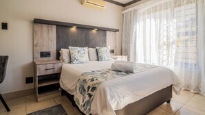 A modern bedroom with a large bed, a wooden nightstand, a bedside table, and a built-in air conditioning unit. - ATKV Goudini Spa (Rawsonville)