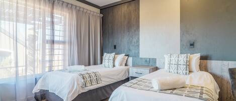 A hotel room with two beds, a ceiling fan, a large window with sheer curtains, and a modern design with a concrete accent wall.