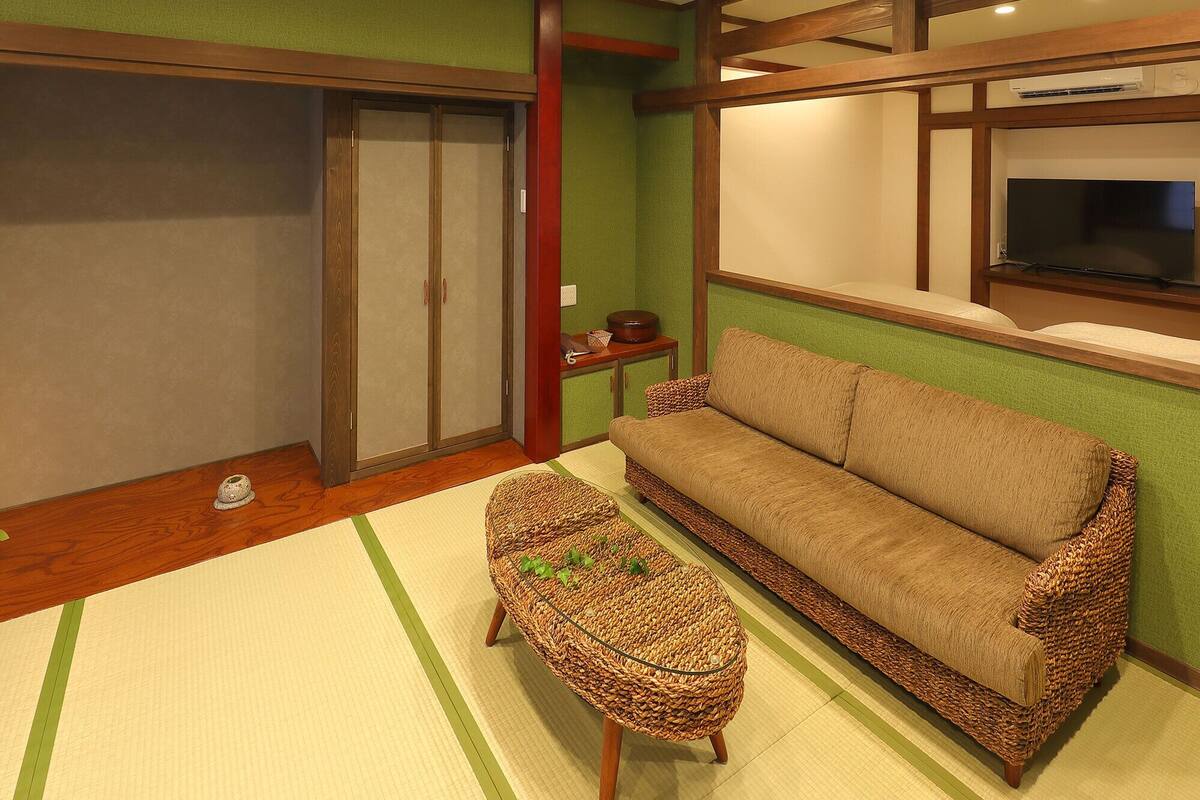 A traditional Japanese-style room with tatami flooring, a low wooden-framed sofa, a woven coffee table, a sliding door, and a mounted television.