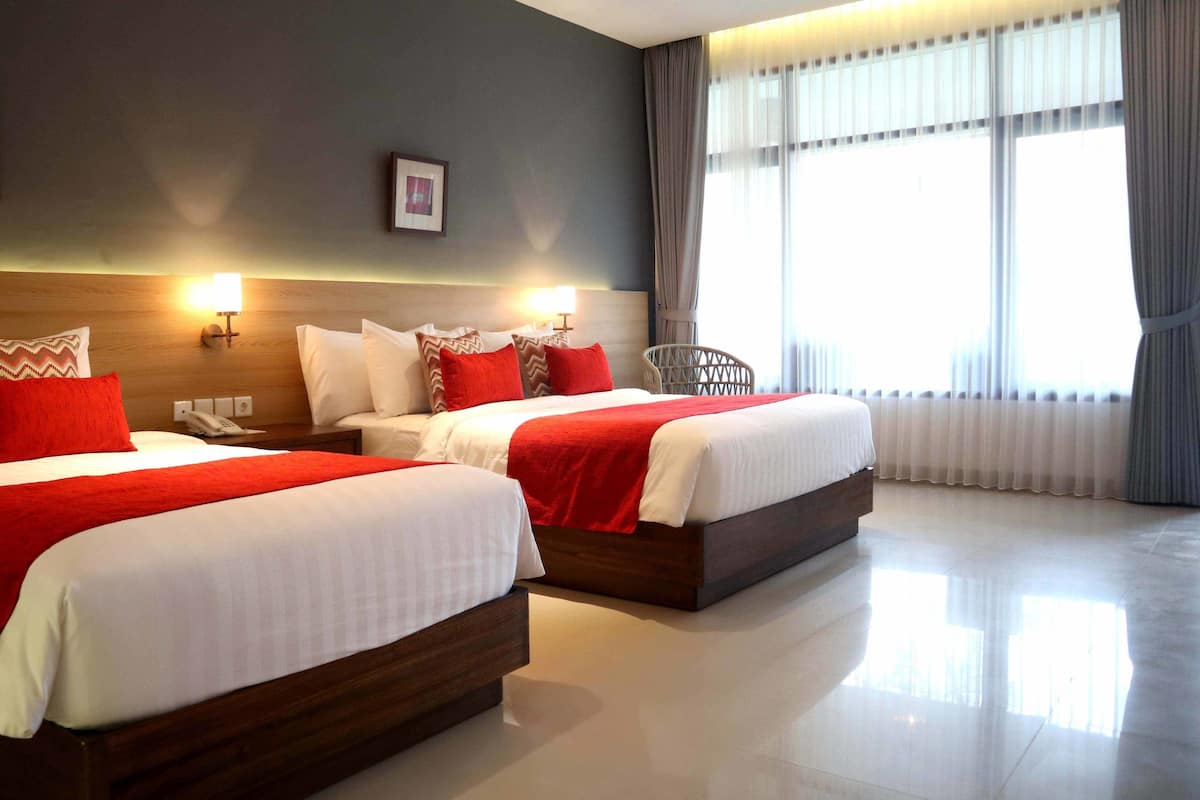 Habitación de hotel con dos camas, cada una con ropa blanca y detalles rojos, un cabecero de madera, una mesita de noche con teléfono y un ventanal con cortinas.