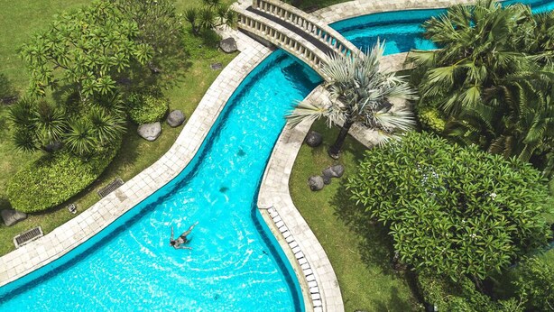 A large, curved swimming pool with a bridge, surrounded by lush greenery and palm trees. - The Cakra Hotel (Denpasar)