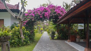 A paved pathway leading through a garden with a wooden pergola covered in pink flowers. - The Cakra Hotel (Denpasar)