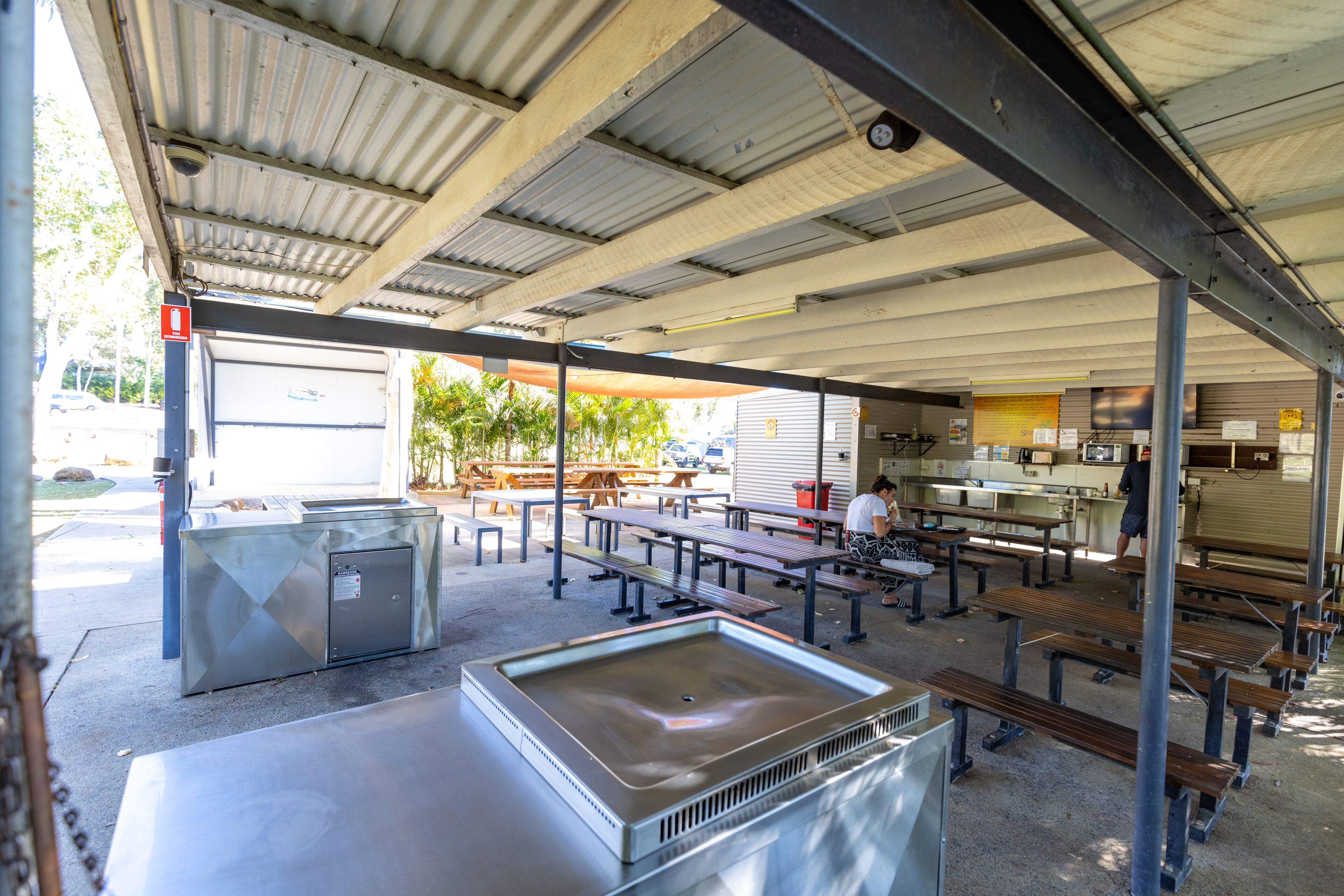 Un espace extérieur aménagé pour les repas, avec des tables et des bancs en métal, un grand congélateur à glaces et une partie abritée où se trouve une cuisine en arrière-plan.