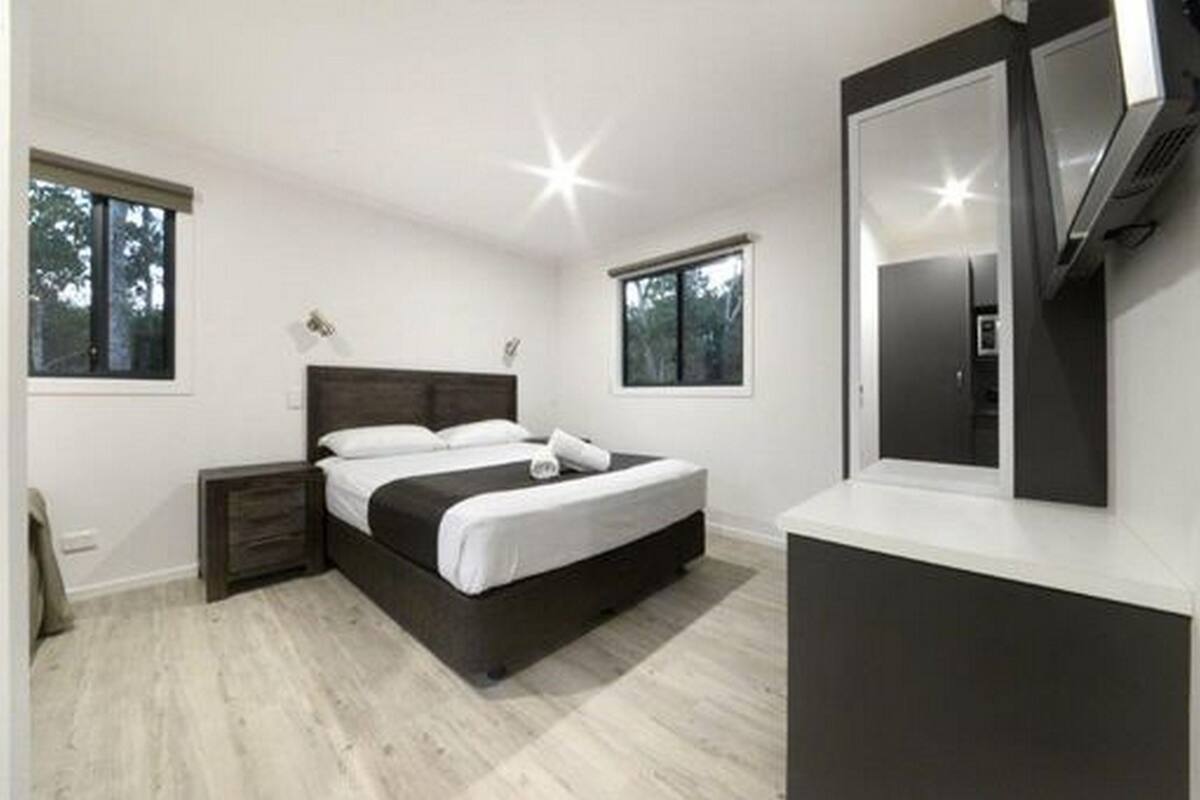Quarto moderno com uma cama grande, mesa de cabeceira, cômoda e espelho. Duas janelas proporcionam luz natural e vista para árvores.