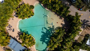 Luchtbeeld van een resort met een zwembad, omringd door palmbomen en een zandstrand met ligbedden.