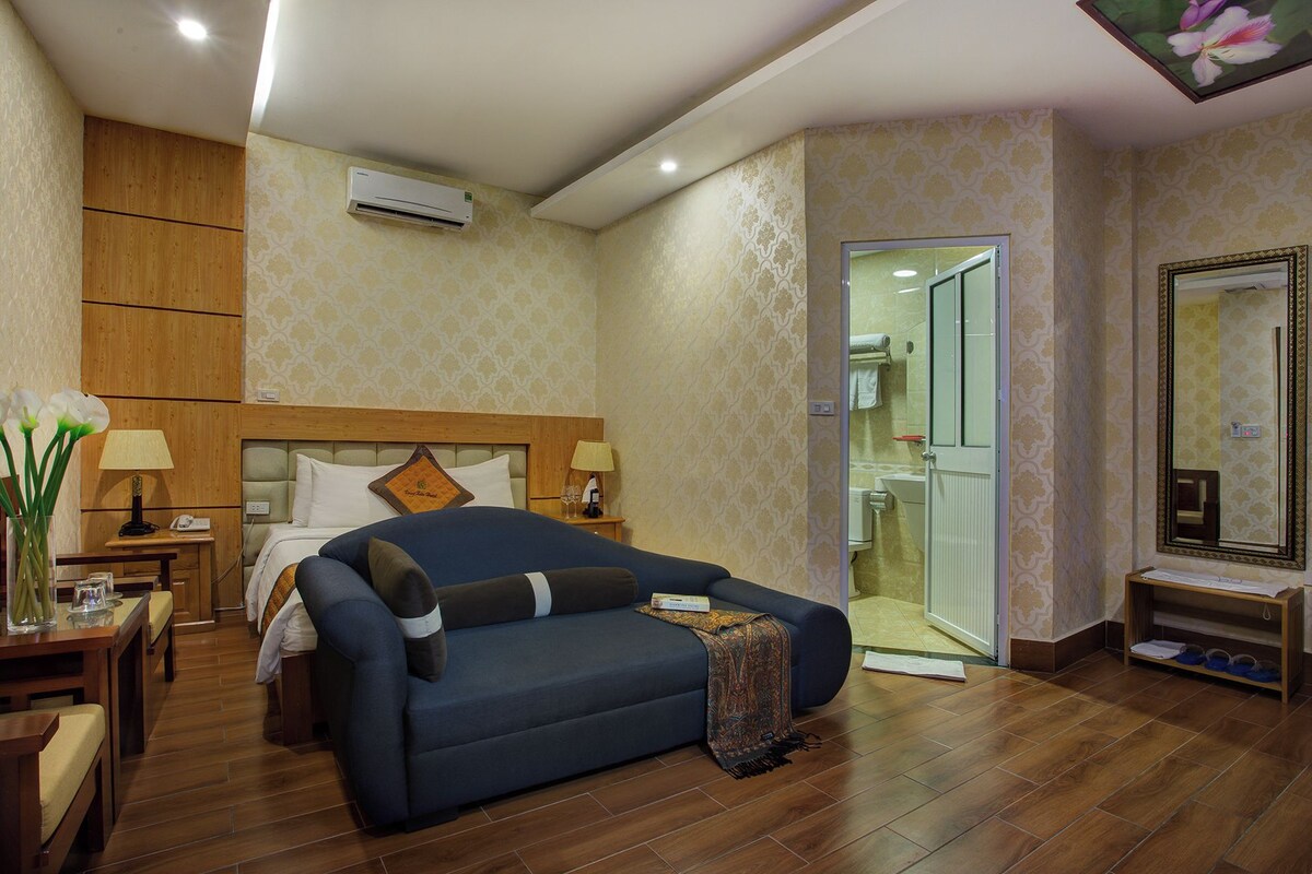 Une chambre d’hôtel avec un lit, un canapé, une table de chevet avec une lampe, un miroir et une salle de bain visible à travers une porte ouverte.