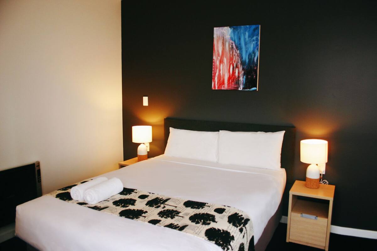 A neatly made bed with white linens, two bedside lamps, and a small wooden nightstand with a lamp.