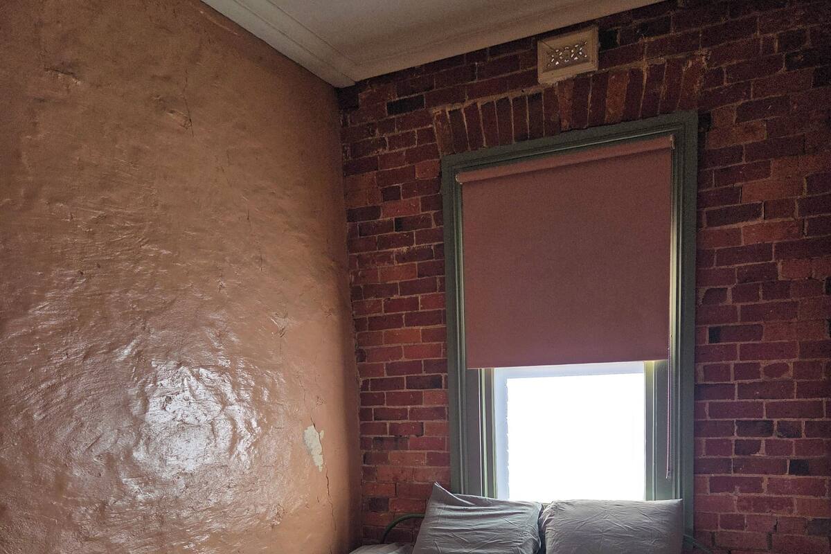 A small, dimly lit room with a single bed, a wooden chair, and a brick wall.