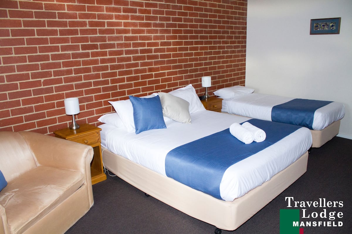 A double room with two beds, each with white and blue bedding, a beige armchair, and a brick wall.