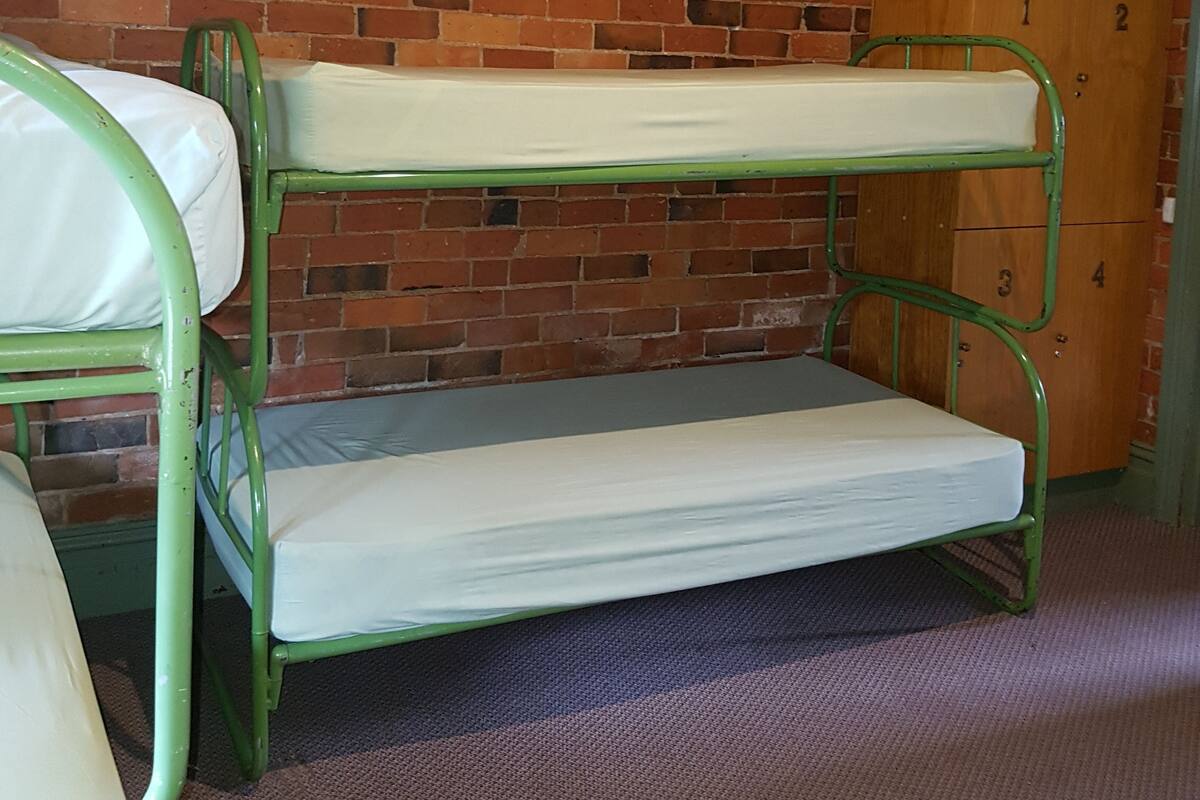 A bunk bed with green metal frames and white bedding in a room with a brick wall and wooden door.