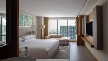 A modern hotel room with a large bed, a sitting area, a TV, and a view of the city.