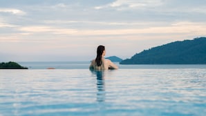 Una persona sentada en una piscina infinita de color azul intenso, con vistas a una isla lejana y un mar tranquilo.