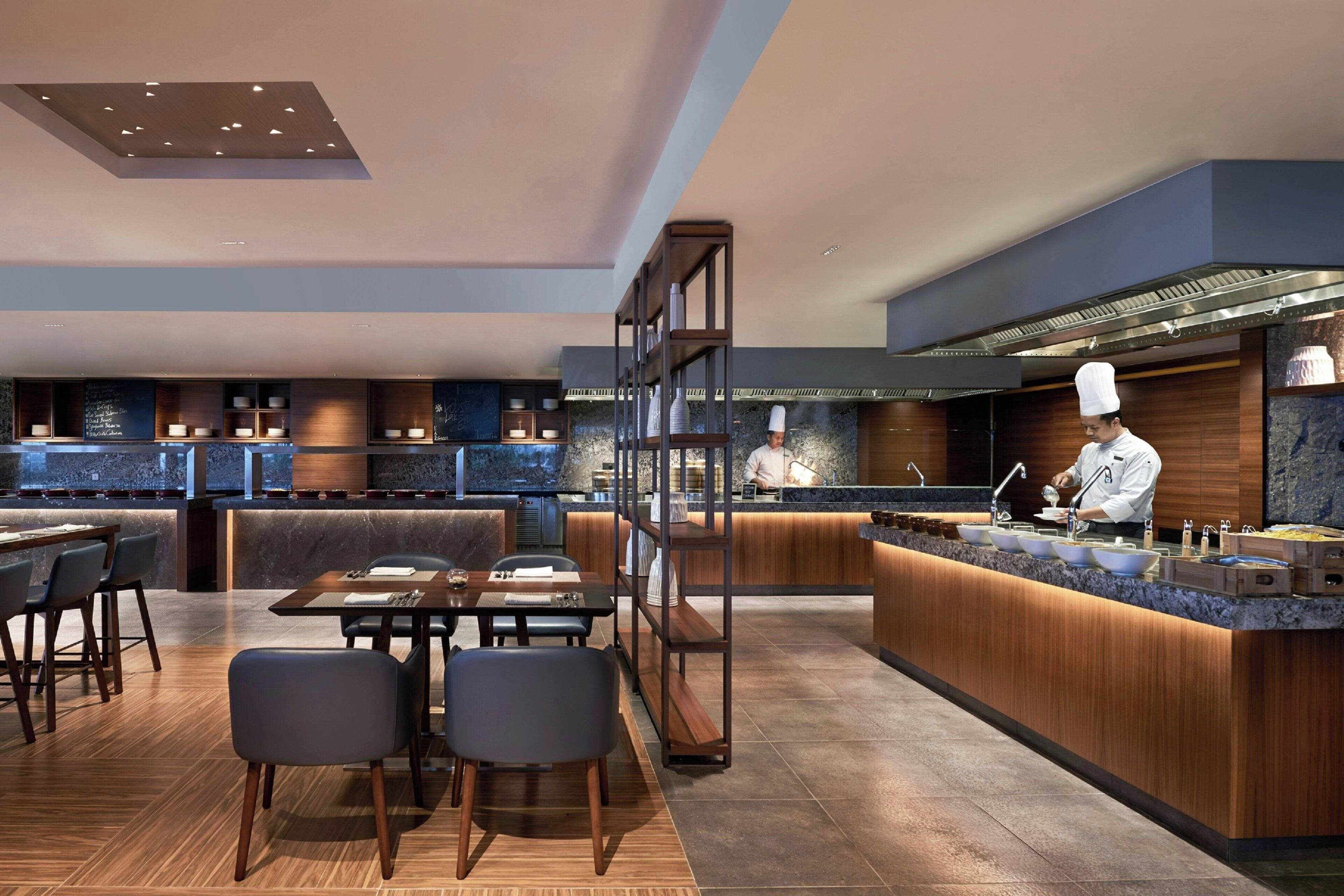 A modern restaurant with a chef at the counter, wooden furniture, and a bar area.
