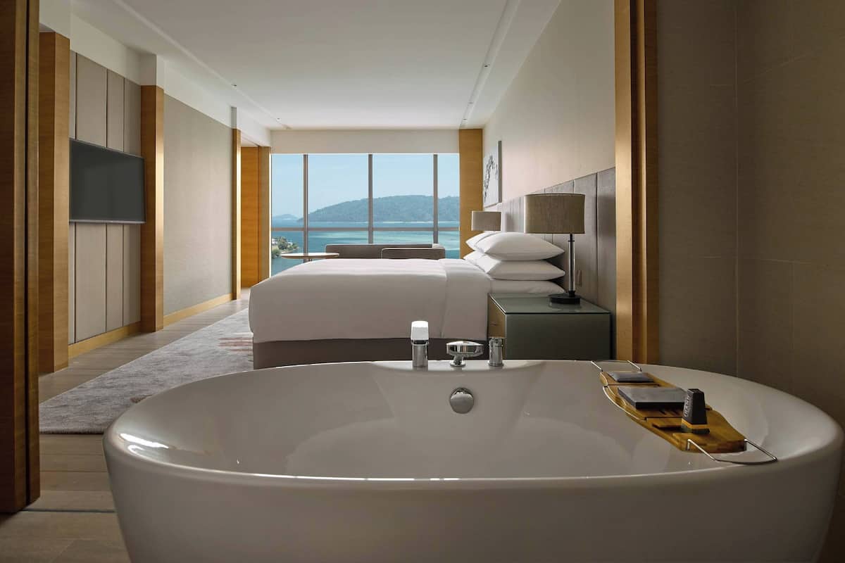 Une chambre d’hôtel moderne dotée d’une grande baignoire, d’un lit avec vue sur la mer et d’une table de chevet avec une lampe.
