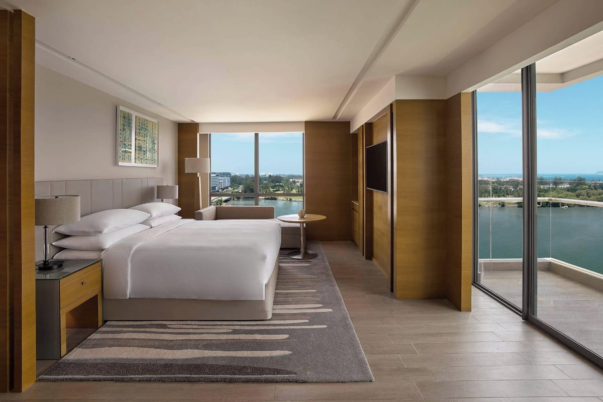 Une chambre d’hôtel moderne, dotée d’un grand lit, d’une table de chevet avec une lampe, d’une petite table ronde et d’un téléviseur. La chambre dispose de grandes fenêtres offrant une vue sur un plan d’eau et des bâtiments.