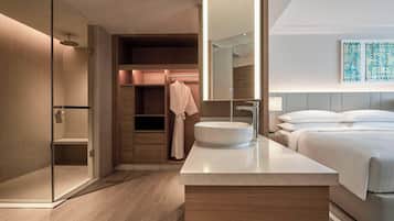 A modern hotel bathroom with a glass-enclosed shower, a large mirror, a round sink, and a bed with white linens.