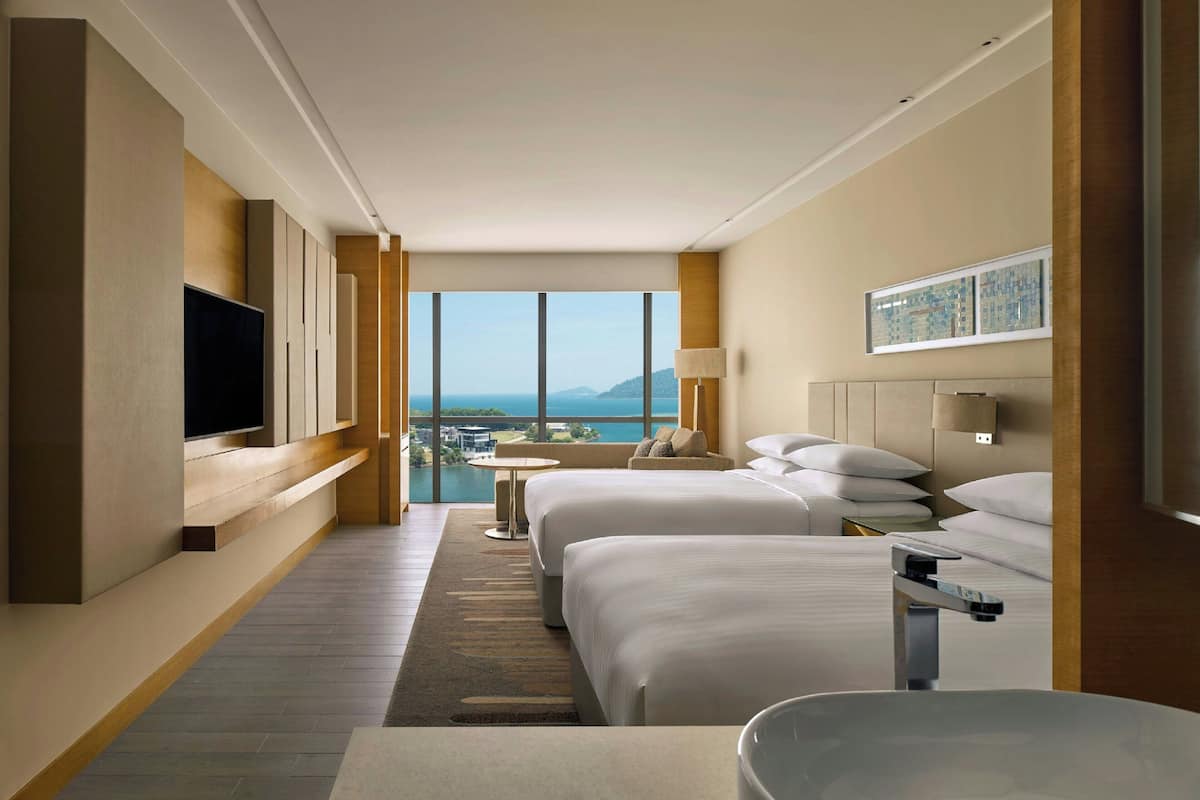 Une chambre d’hôtel avec deux lits, une télévision, une vue sur la mer et une petite table avec une lampe.
