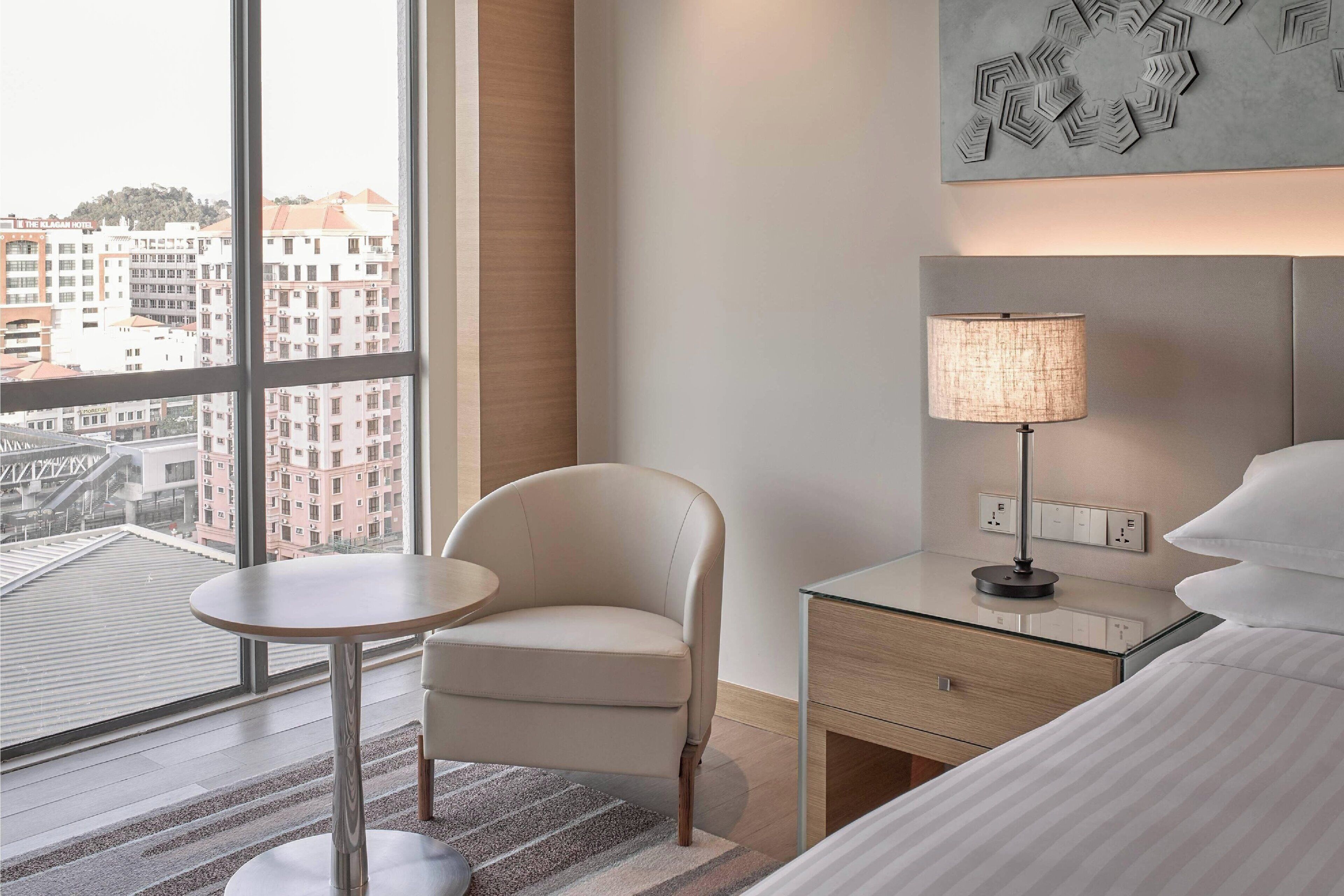 Quarto de hotel com uma janela grande, uma mesa redonda, uma cadeira, um abajur, uma cama e uma obra de arte na parede.
