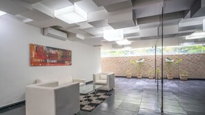 Um lobby moderno com uma mesa de centro de vidro, sofás brancos, uma grande pintura abstrata e plantas em vasos, tudo frente a uma parede de tijolos.