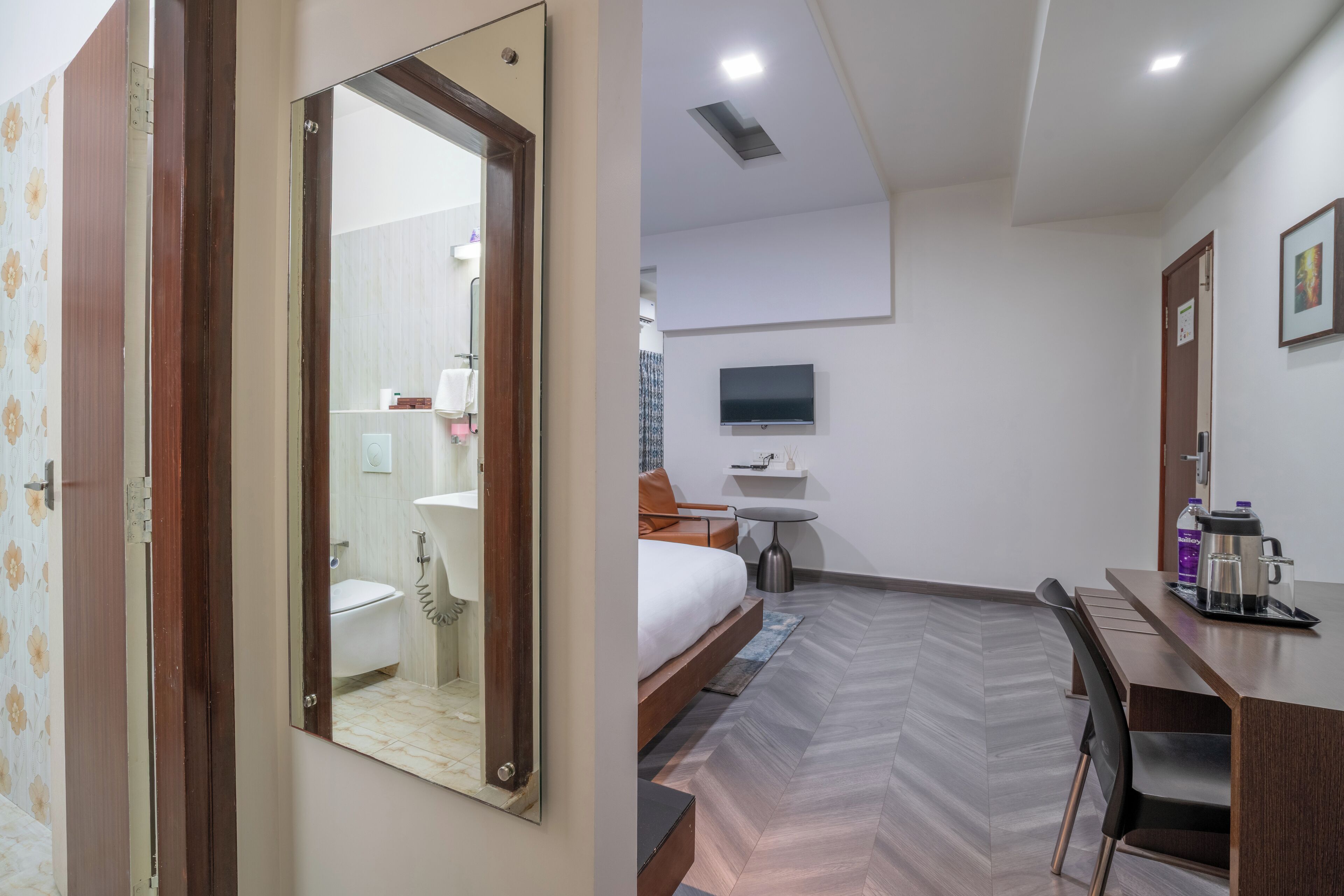A hotel room with a bed, a desk, a chair, a TV, a bathroom, and a mirror.