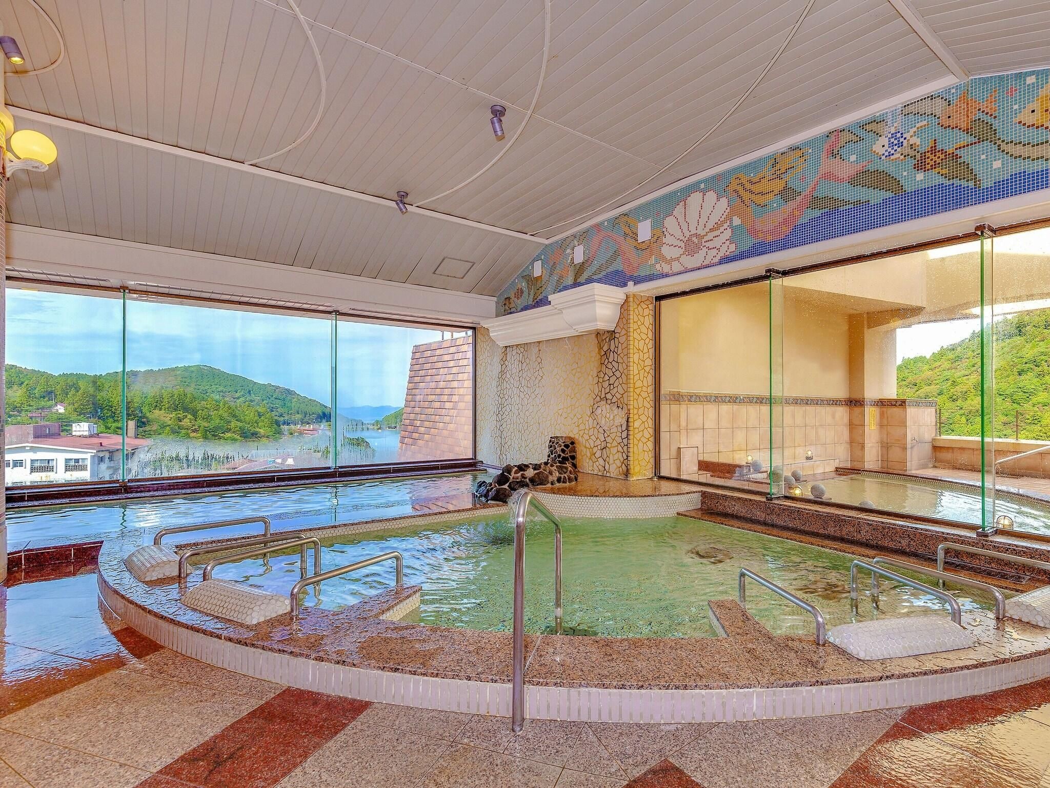 An indoor swimming pool with a view of a lake and hills, surrounded by a tiled floor and a mosaic wall.