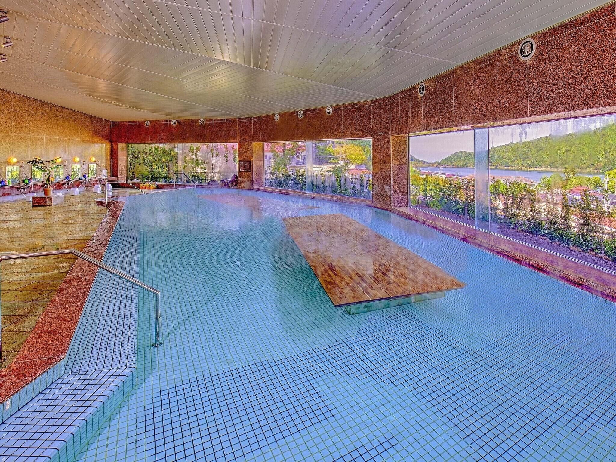 An indoor swimming pool with a wooden deck, tiled floor, and large windows offering a view of greenery and buildings.